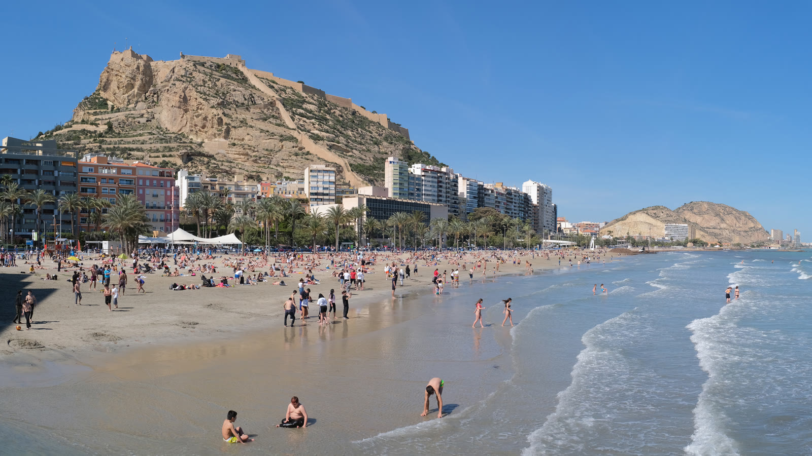 Postiguet-stranden i Alicante med badegæster i vandkanten og borgen Castillo de Santa Bárbara på klippen bag byen