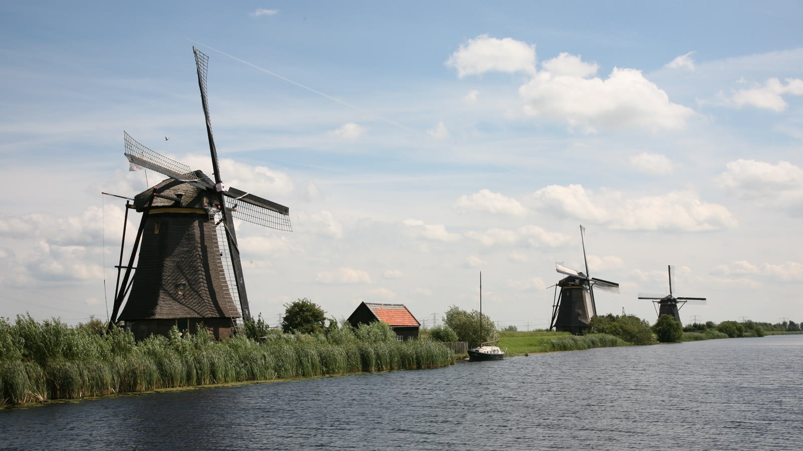 Tre vindmøller langs kanalen ved Kinderdijk med siv i forgrunden og blå himmel