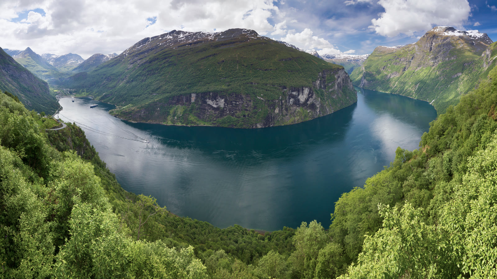 Geirangerfjorden set fra Ørnesvingen med grøn fjeldryg, snedækkede tinder og bygden Geiranger i bunden af fjorden